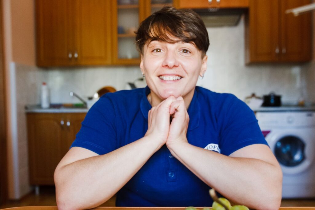 A cheerful woman in a blue shirt smiling while seated in a cozy kitchen environment.