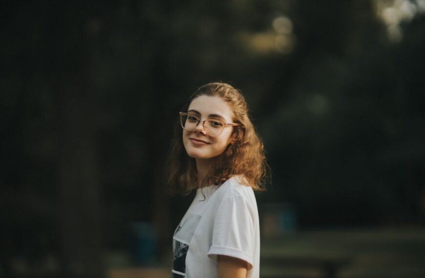 A young woman smiling warmly outdoors, wearing glasses and a white shirt.