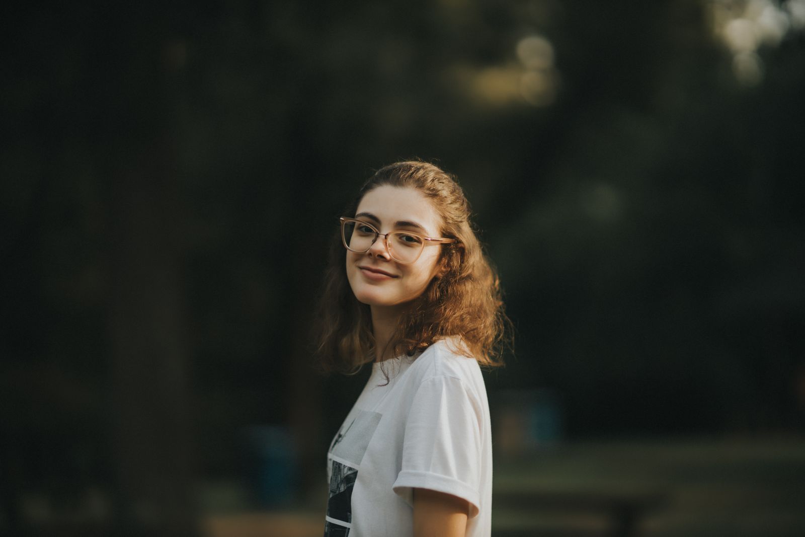 A young woman smiling warmly outdoors, wearing glasses and a white shirt.
