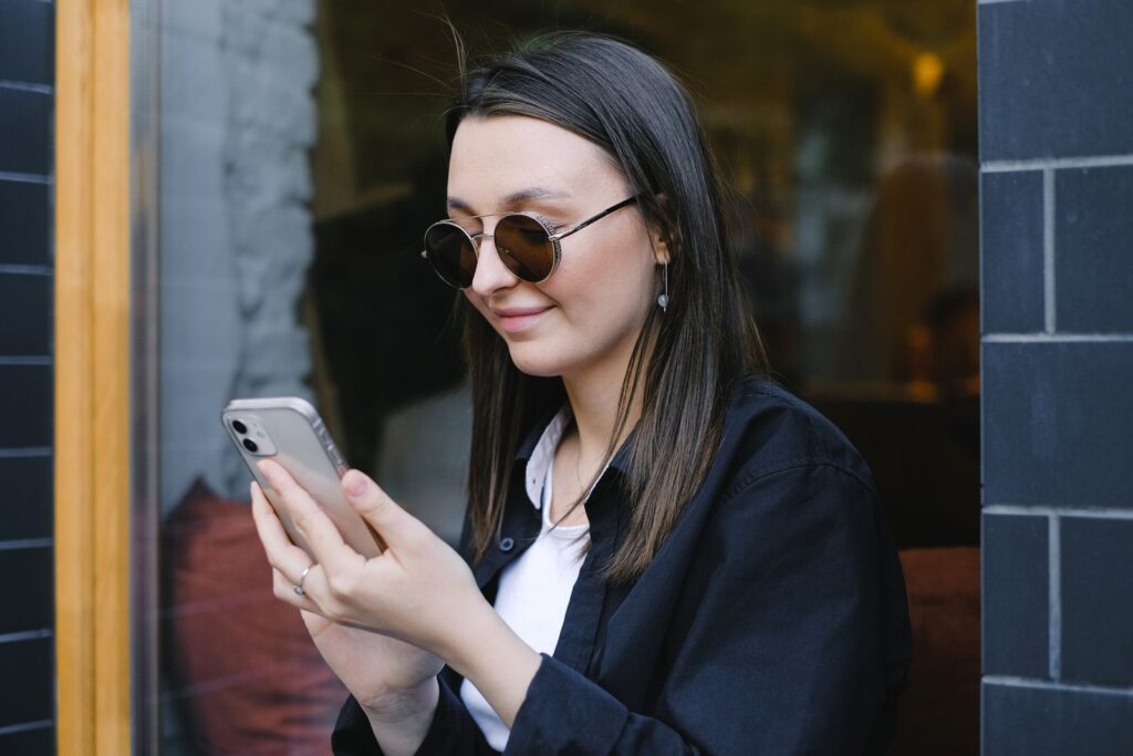 A young woman with sunglasses smiles while using her smartphone outdoors.