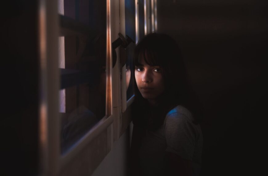 A woman leans by a window in a dark room, illuminated by soft natural light.