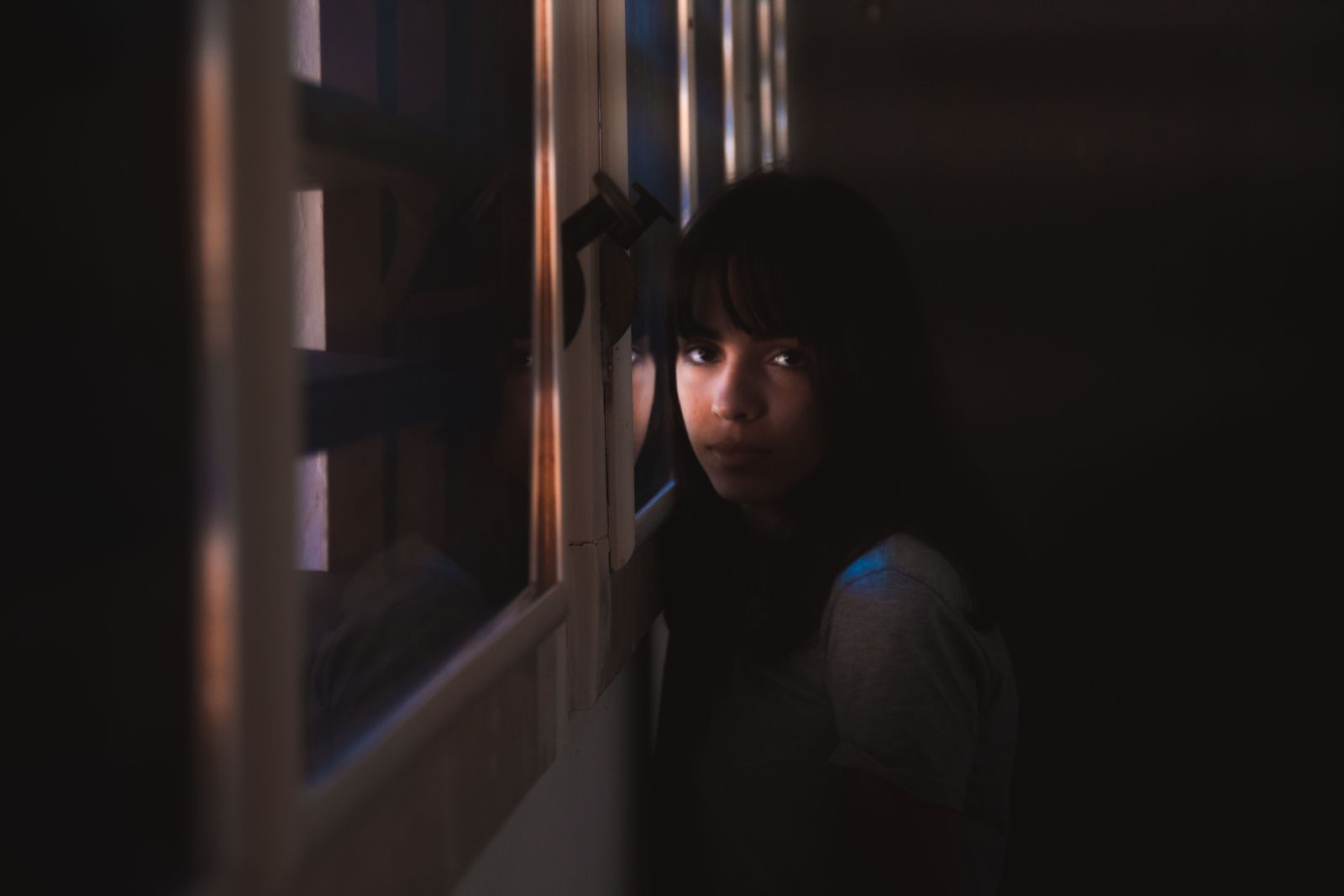 A woman leans by a window in a dark room, illuminated by soft natural light.