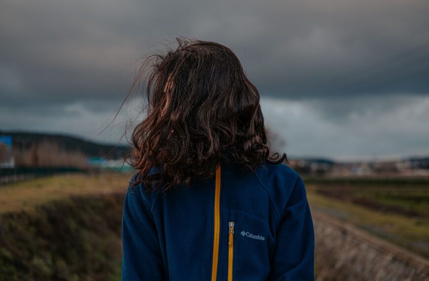 A woman stands outdoors wearing a jacket, facing away, with storm clouds overhead.