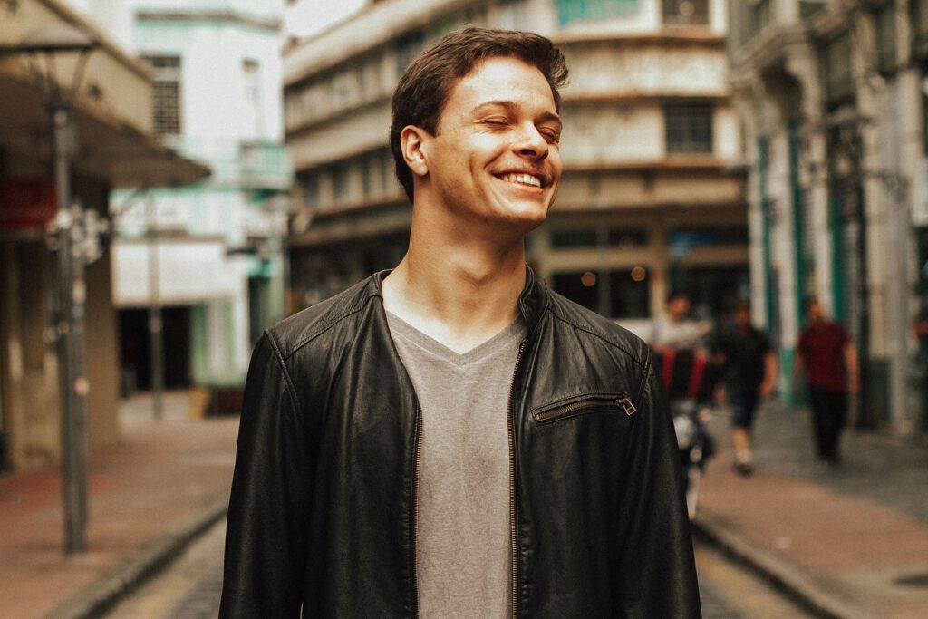 A young man smiling confidently in a city street wearing a leather jacket, exuding urban fashion style.