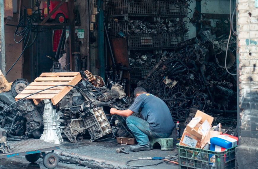 Mechanic fixing auto parts in a cluttered urban workshop, surrounded by machinery.