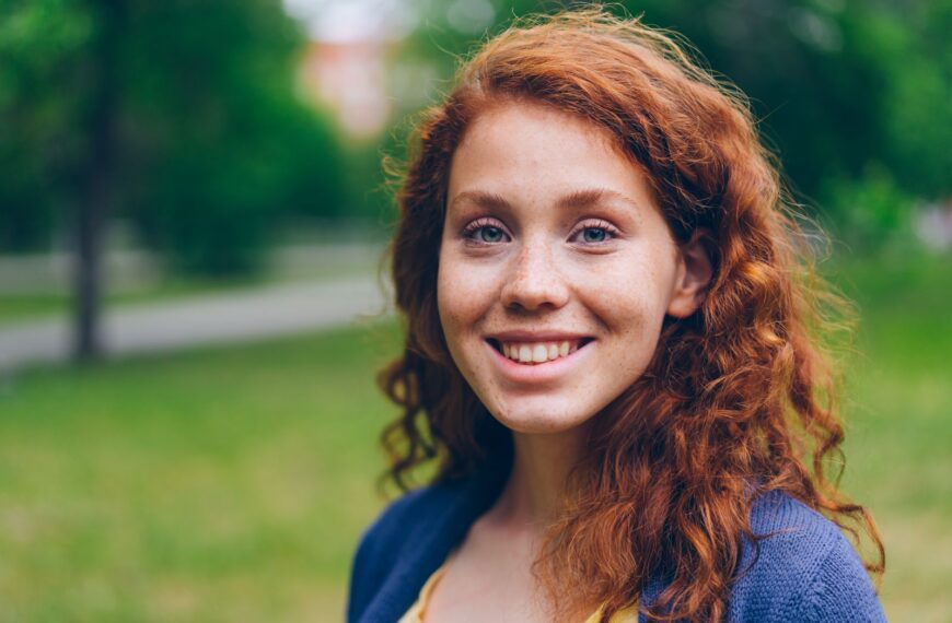 Portrait of a cheerful young woman with red hair in a green outdoor setting.