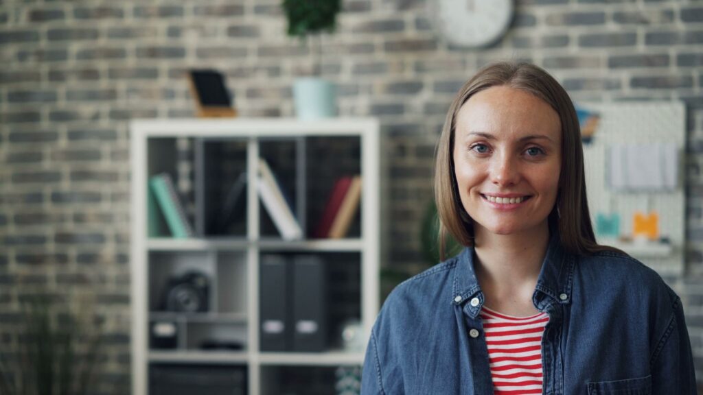 Portrait of a smiling woman wearing a jean shirt in a modern office with brick wall background.