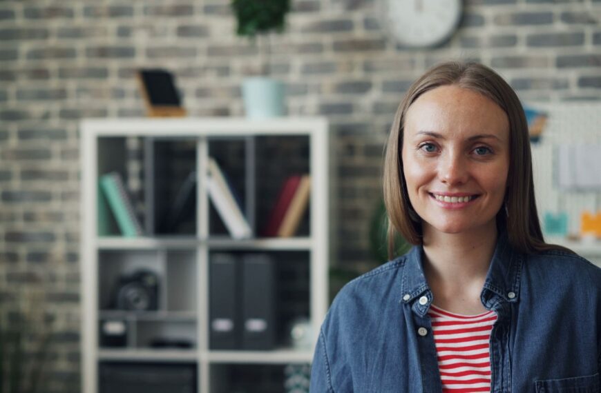 Portrait of a smiling woman wearing a jean shirt in a modern office with brick wall background.