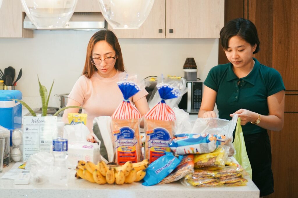 Two women unpack groceries in a modern kitchen setting, featuring bread, bananas, and a variety of packaged foods.