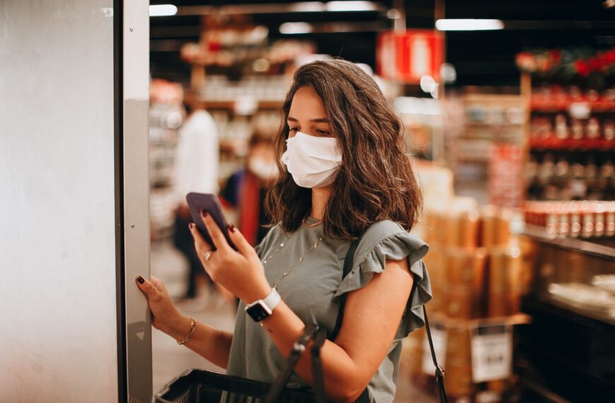 Woman wearing mask shopping in a supermarket, checking smartphone. Captures modern retail experience.