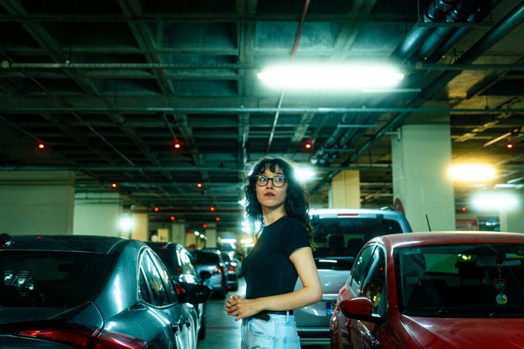 Stylish woman standing in a dimly lit urban parking garage surrounded by parked cars.