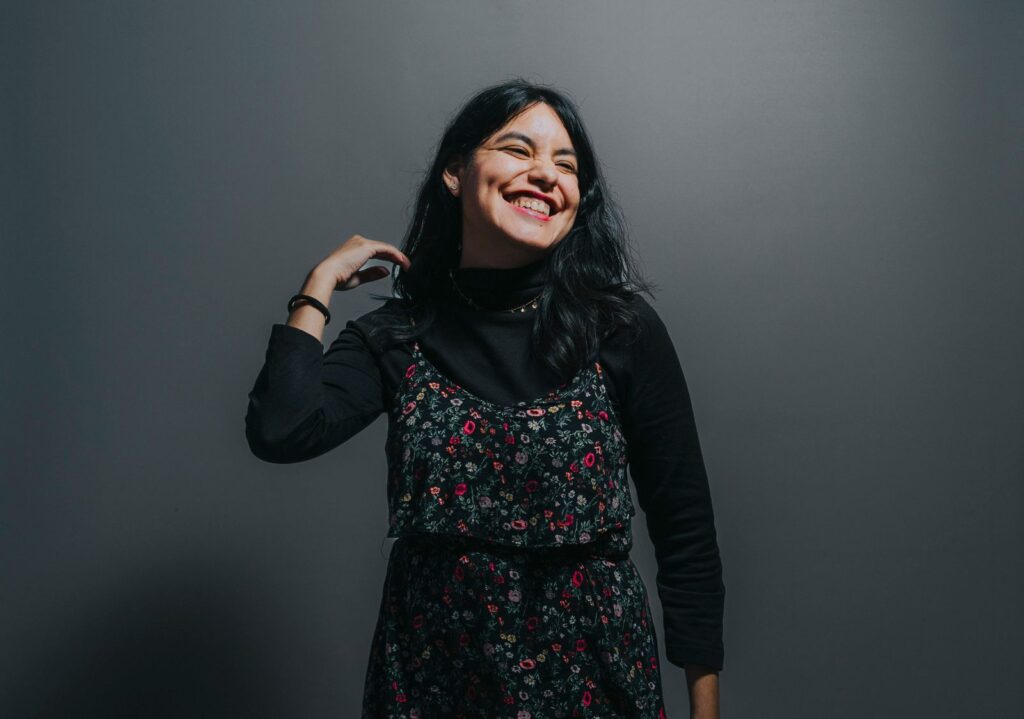 Smiling woman posing confidently in a floral dress against a grey background, exuding joy and positivity.