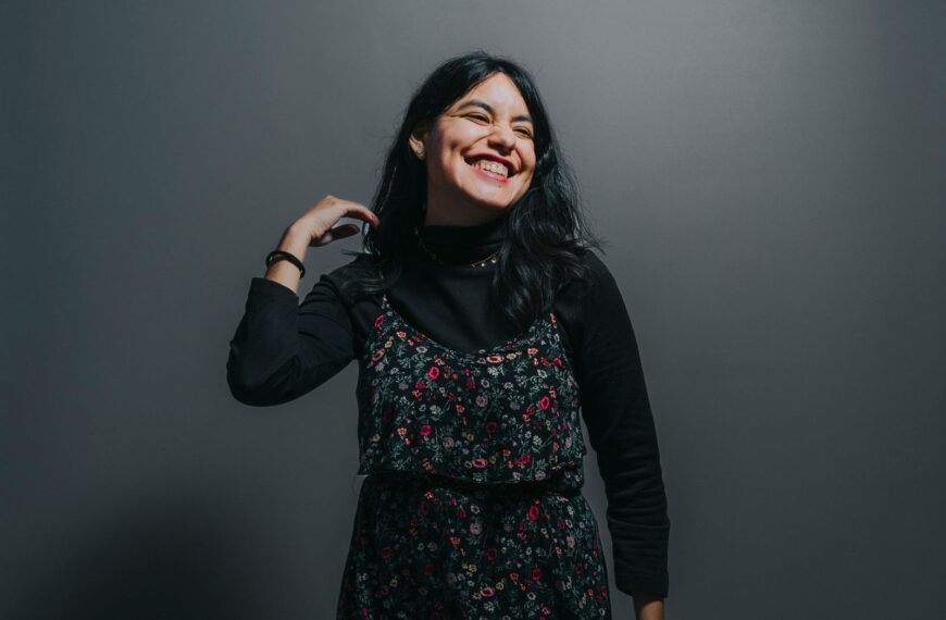 Smiling woman posing confidently in a floral dress against a grey background, exuding joy and positivity.