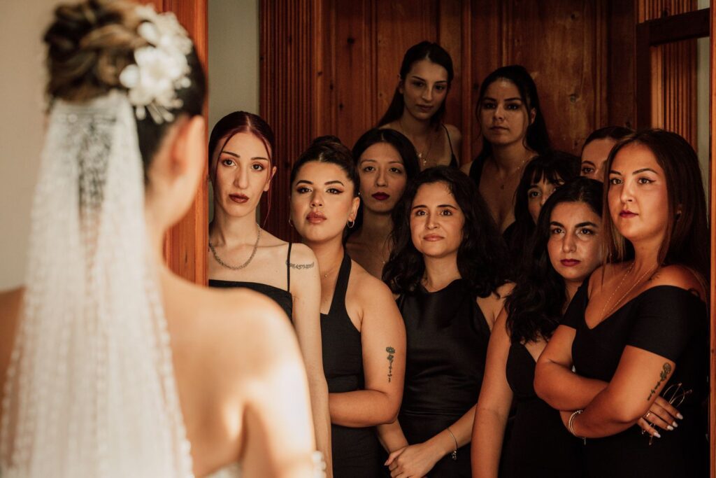 The bride faces a group of bridesmaids in elegant attire, creating a warm indoor atmosphere.