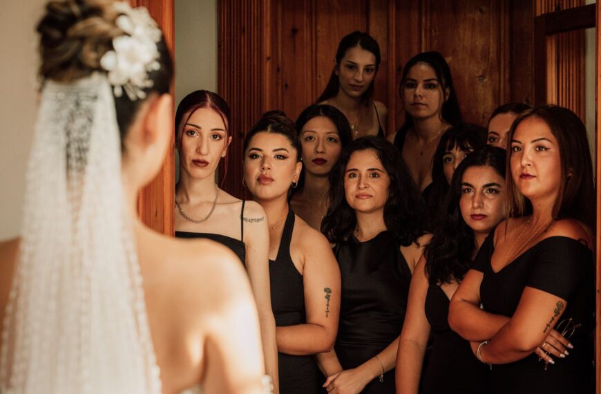 The bride faces a group of bridesmaids in elegant attire, creating a warm indoor atmosphere.