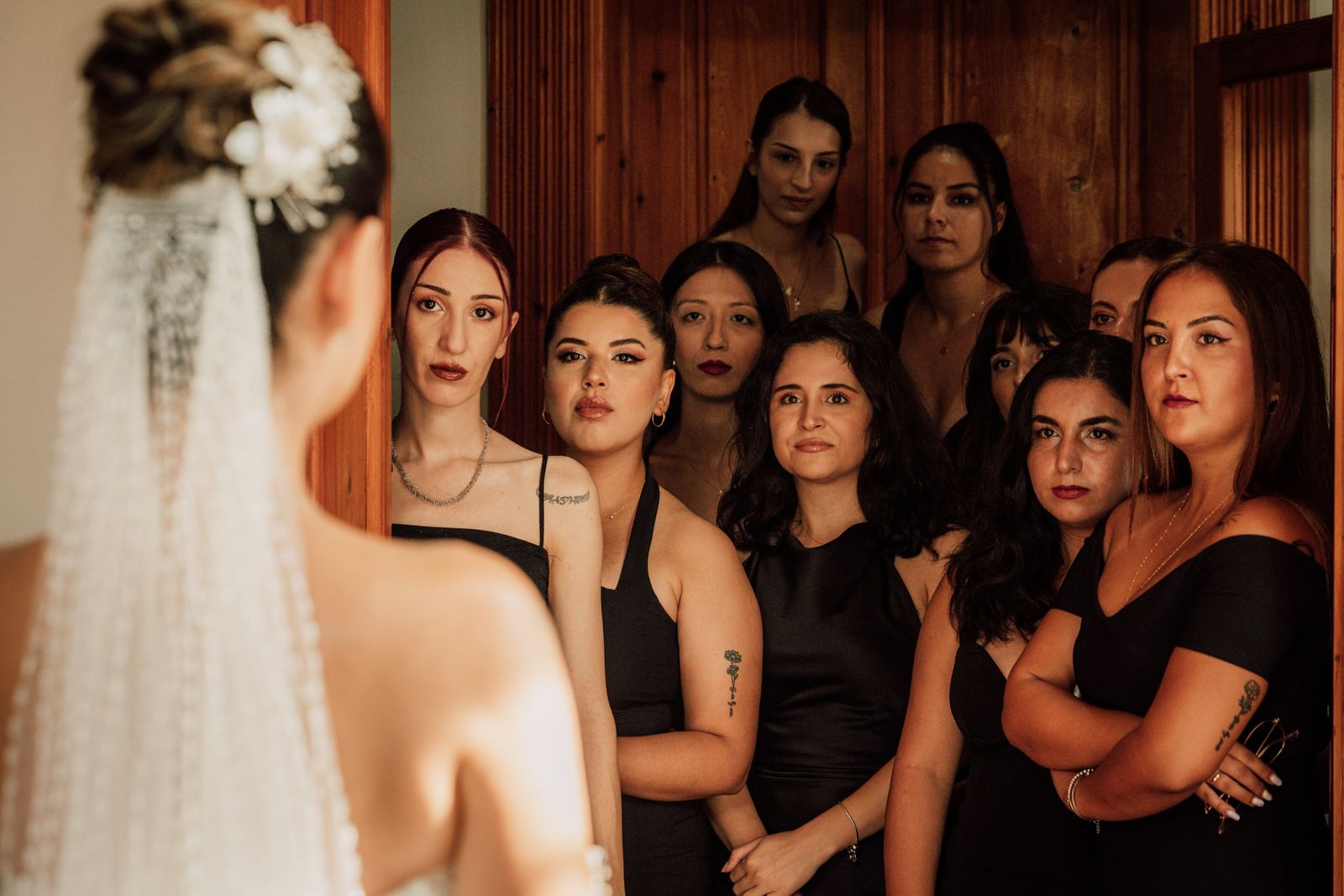 The bride faces a group of bridesmaids in elegant attire, creating a warm indoor atmosphere.