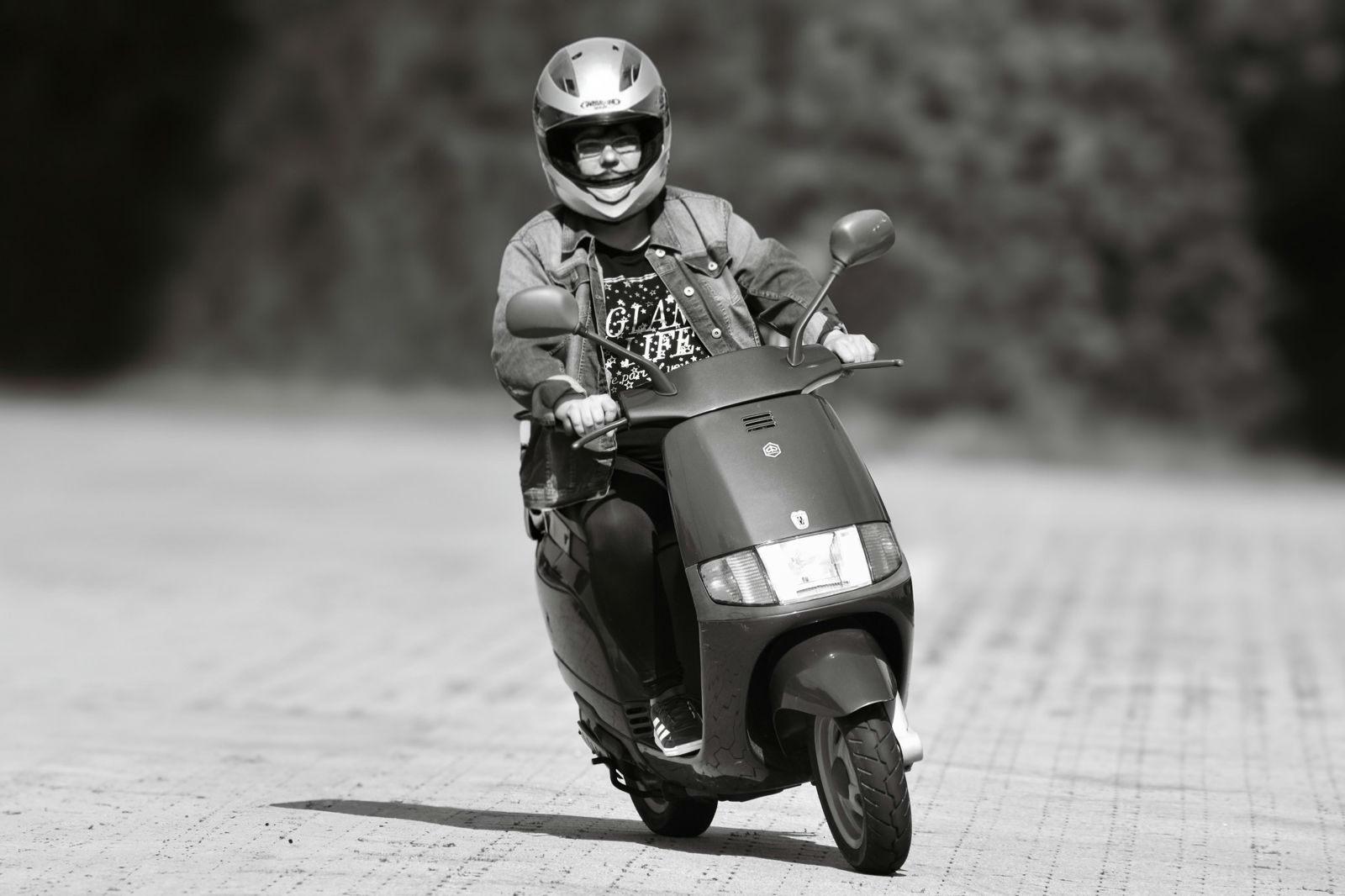 A person rides a scooter wearing protective gear on an outdoor path in black and white photo.