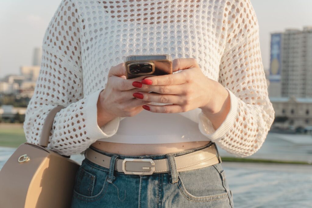 Close-up of a woman using a smartphone outdoors, wearing a stylish casual outfit with red nails.