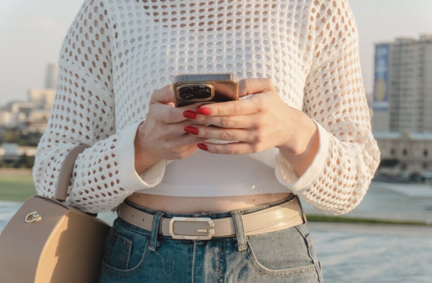 Close-up of a woman using a smartphone outdoors, wearing a stylish casual outfit with red nails.