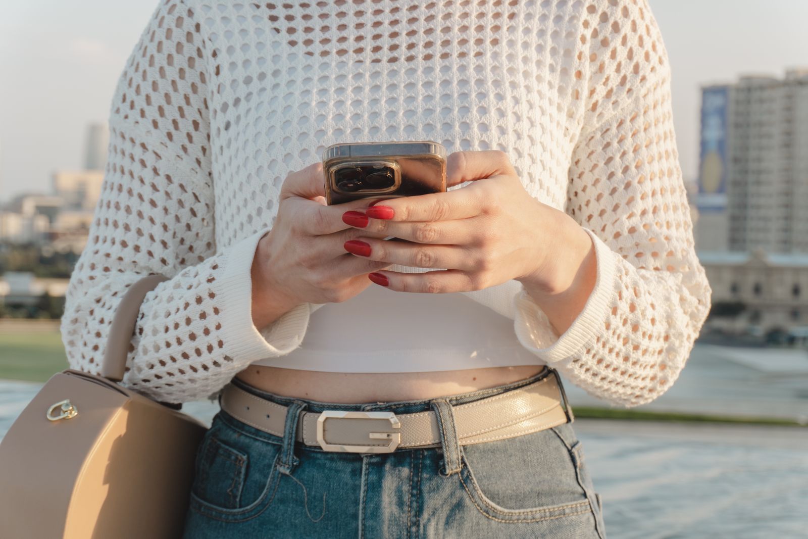 Close-up of a woman using a smartphone outdoors, wearing a stylish casual outfit with red nails.