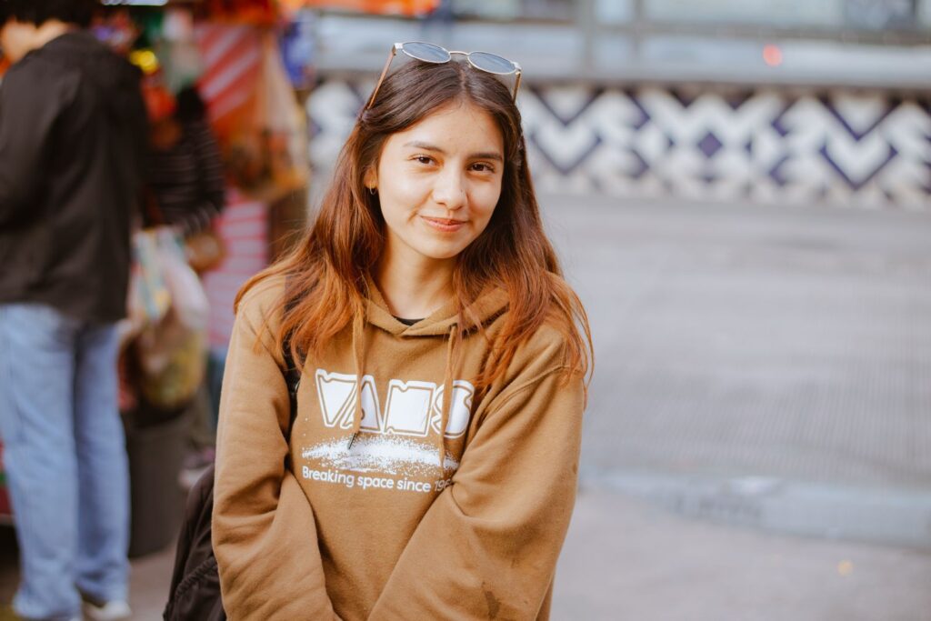 A young woman smiles in a lively urban setting, donning a casual hoodie.