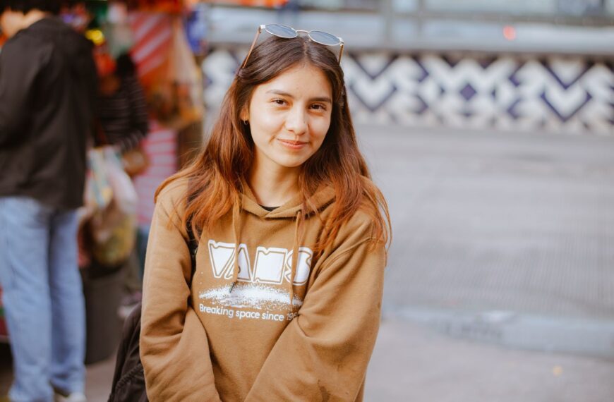 A young woman smiles in a lively urban setting, donning a casual hoodie.