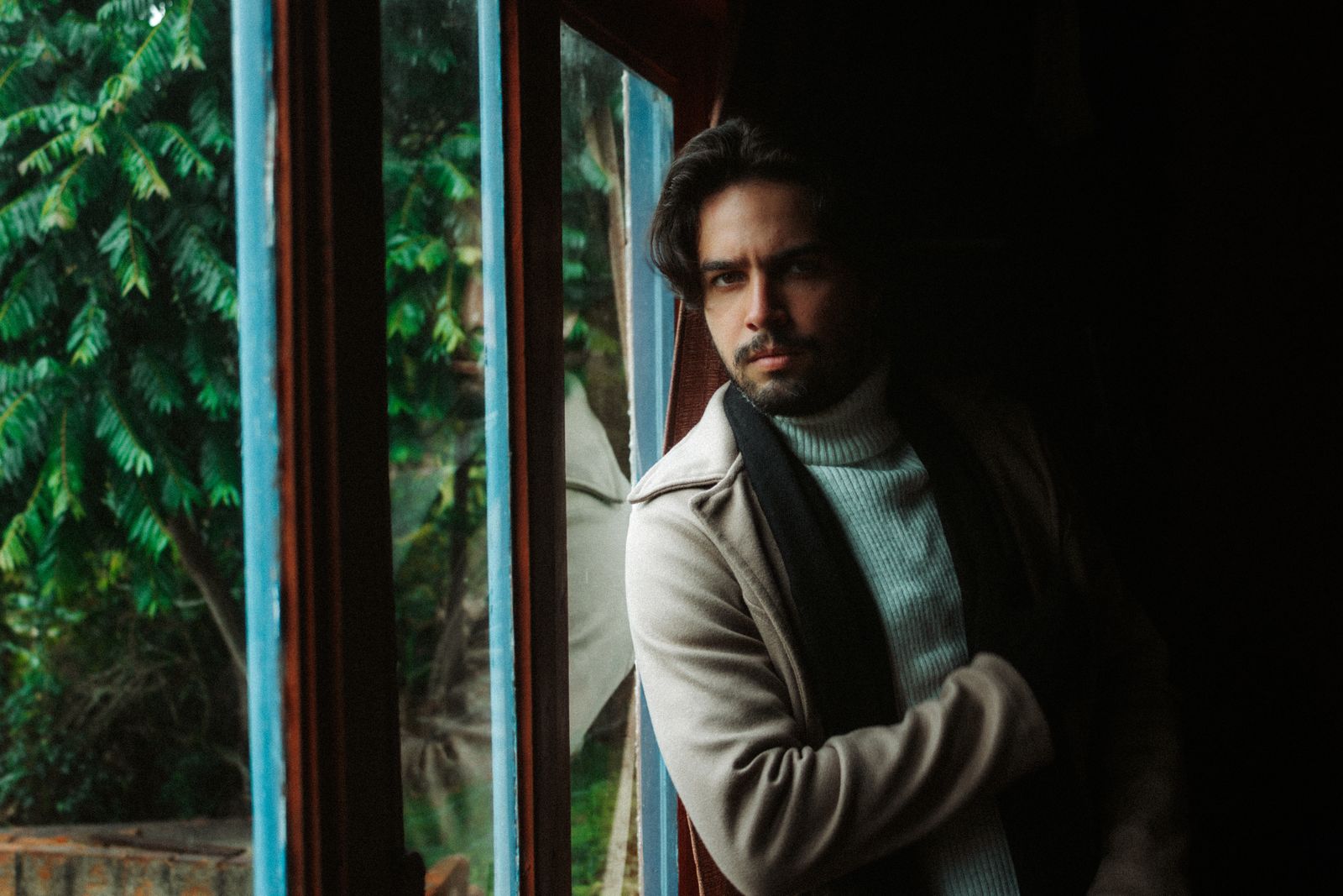 A thoughtful man gazes out a window, wearing a cozy sweater and coat indoors.