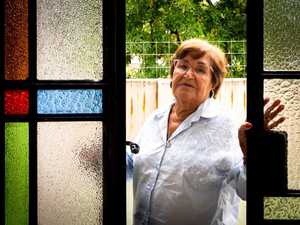 Elderly woman in white shirt poses by colorful stained glass door with a thoughtful expression.