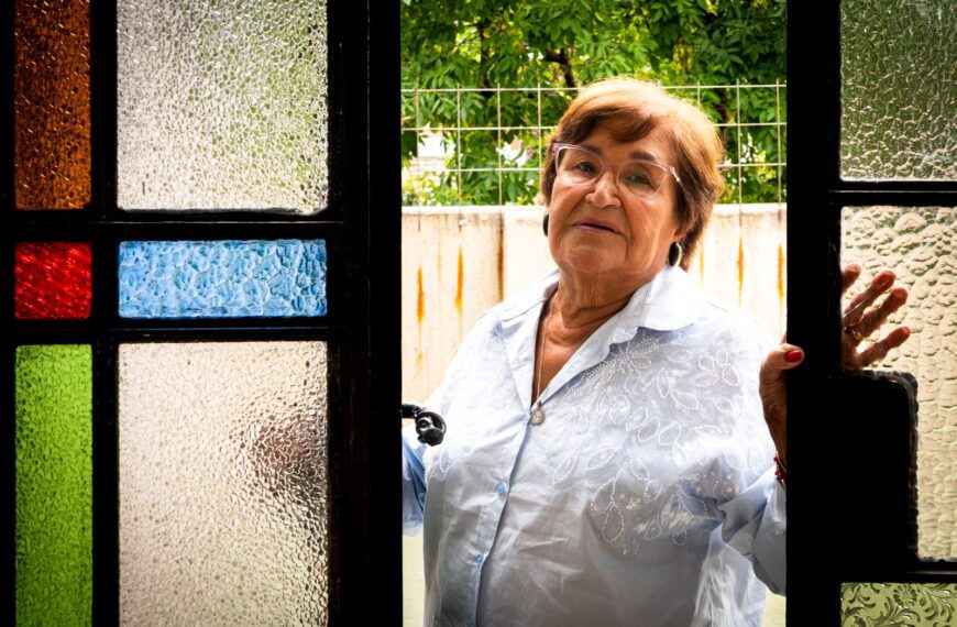 Elderly woman in white shirt poses by colorful stained glass door with a thoughtful expression.