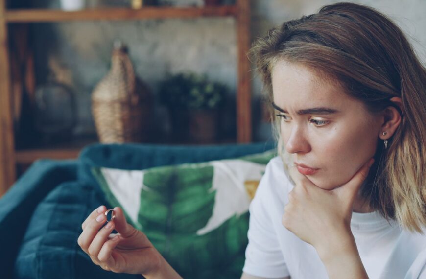 Young woman sitting indoors, holding a ring thoughtfully, looking pensive.