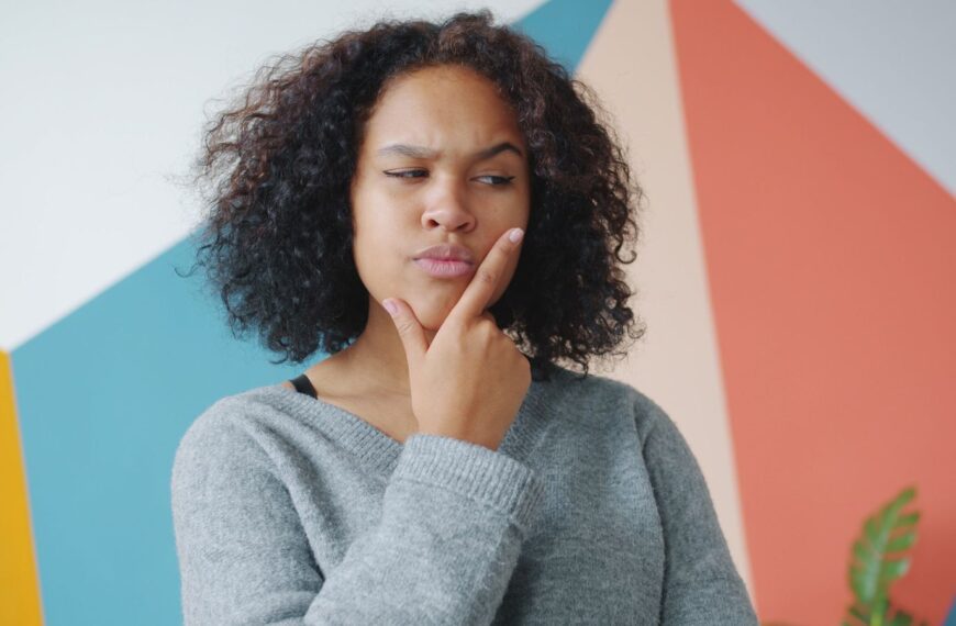 Pensive woman with curly hair pondering in front of colorful geometric wall.