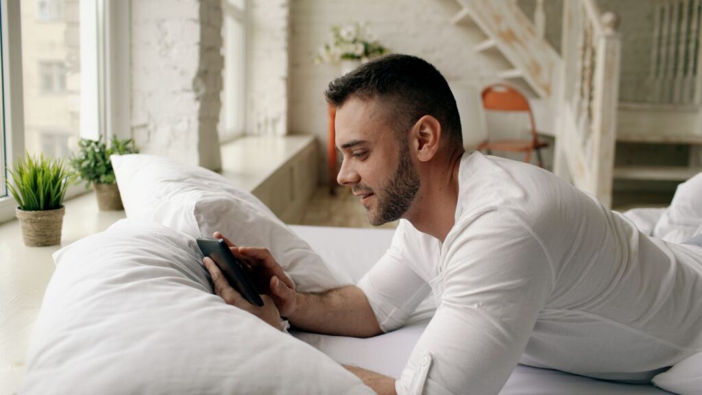Adult male relaxing on bed using smartphone in modern, bright room.