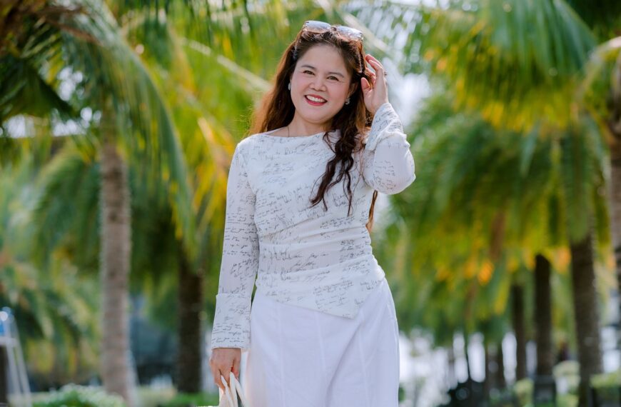 Smiling woman in white outfit walking among vibrant green palm trees on a sunny day.