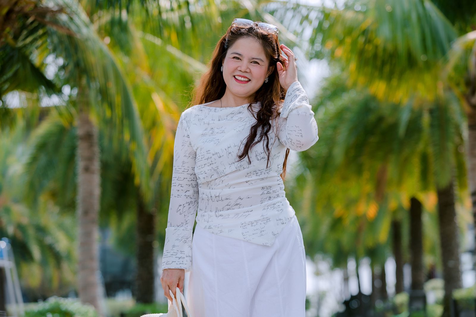 Smiling woman in white outfit walking among vibrant green palm trees on a sunny day.