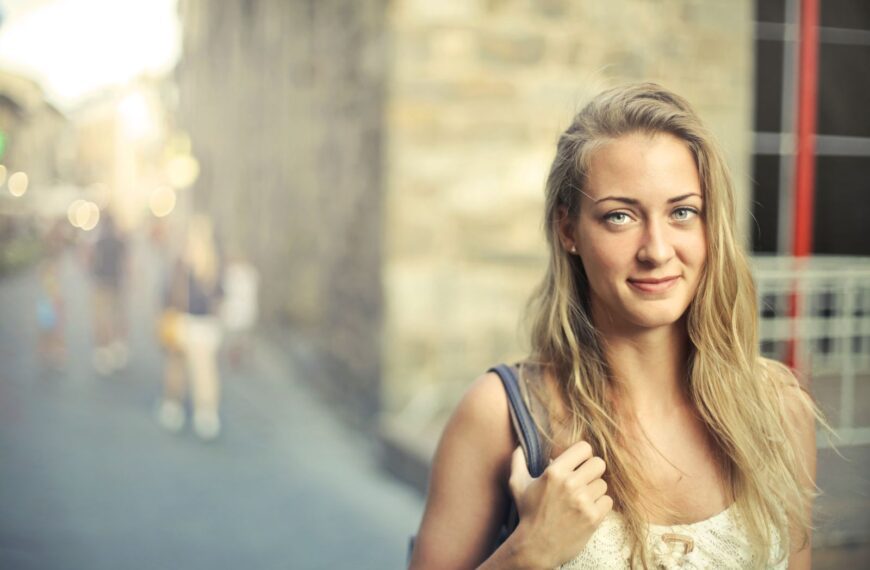 Portrait of a smiling blonde woman carrying a bag, with a bright and cheerful expression in an urban setting.