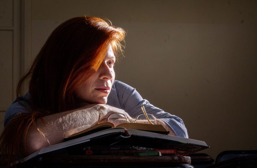 A thoughtful student sits with books in a classroom, illuminated by natural light.