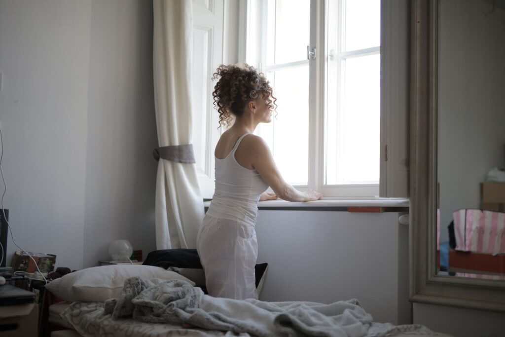 Back view of curly hair female wearing pajamas standing on bed and looking out of window after awakening in bedroom