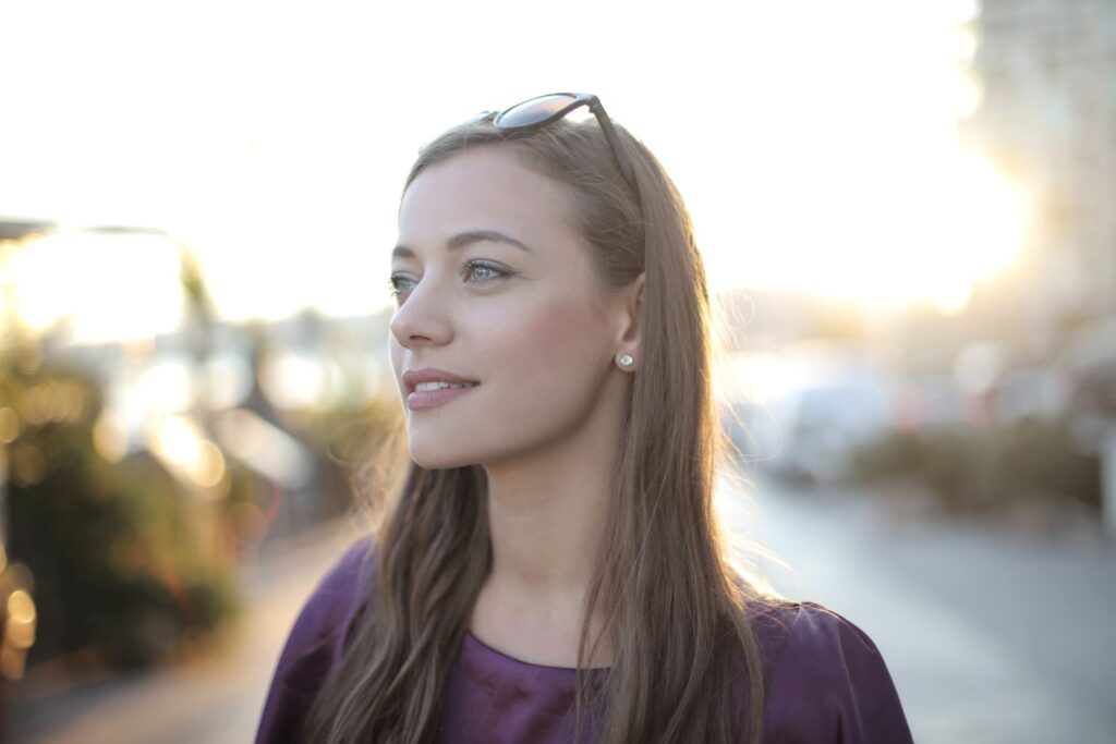 A woman with long hair and a pensive expression smiles softly in an outdoor setting at sunset.
