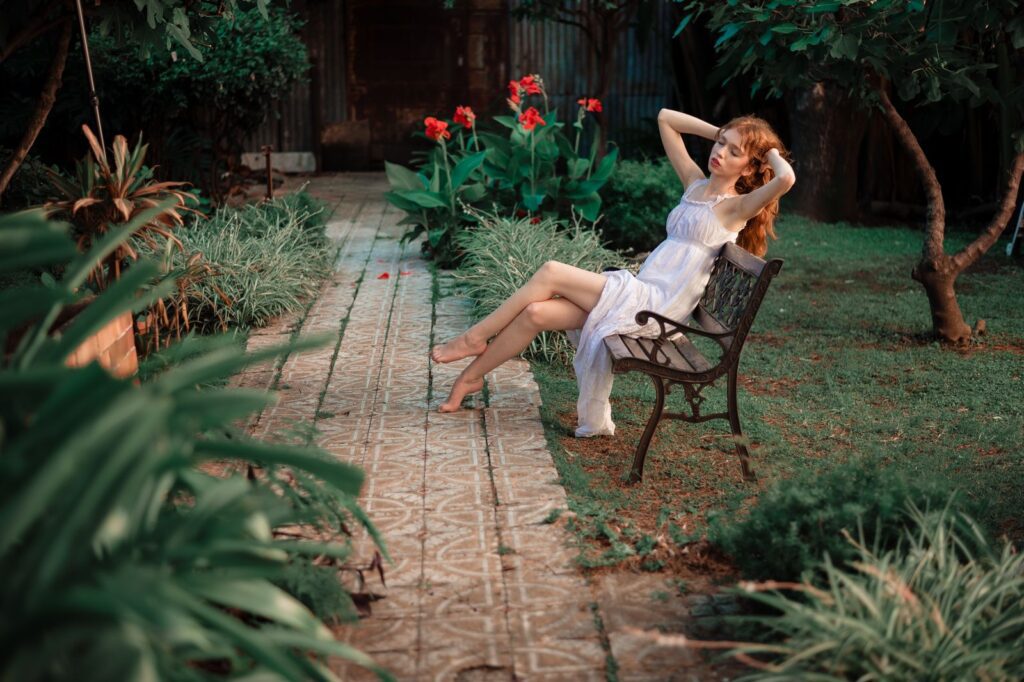 Serene portrait of a woman in a white dress relaxing on a garden bench surrounded by lush greenery and vibrant flowers.