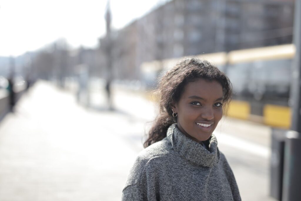Young woman smiling on a sunny day in an urban setting, wearing a cozy sweater.