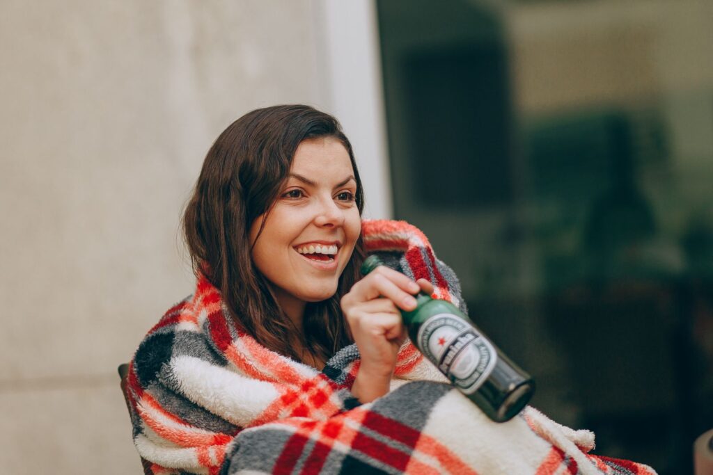 A cheerful adult wrapped in a plaid blanket, enjoying a beer outdoors, radiating happiness.
