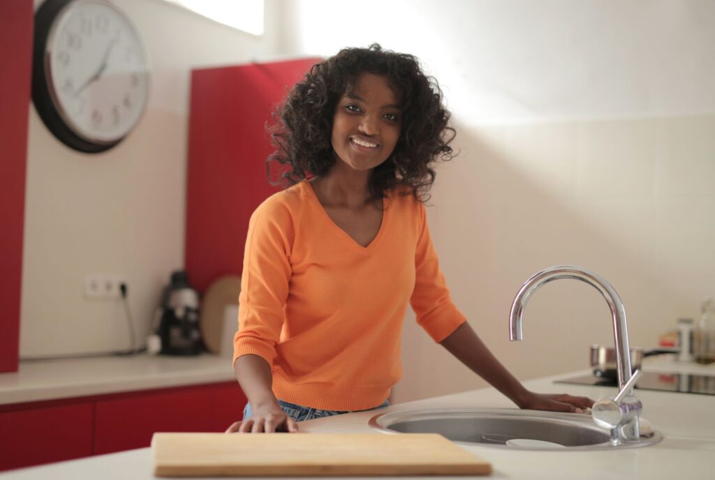 Smiling African American woman in casual wear standing at countertop by sink and cutting board placed aside in cozy kitchen and looking at camera