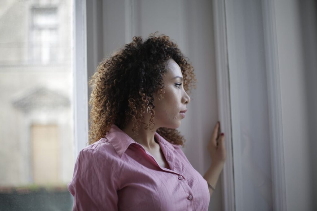 Pensive young ethnic lady in casual pink shirt standing near window and looking away thoughtfully while resting in modern light apartment