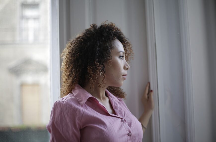 Pensive young ethnic lady in casual pink shirt standing near window and looking away thoughtfully while resting in modern light apartment