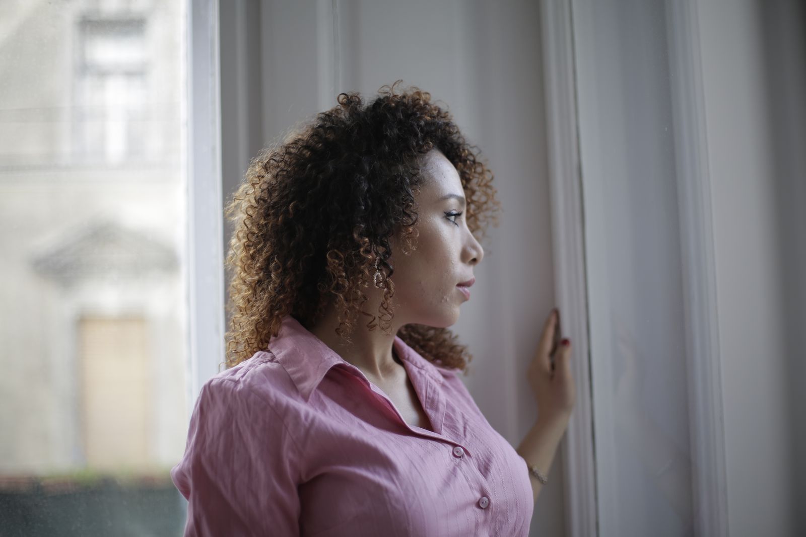 Pensive young ethnic lady in casual pink shirt standing near window and looking away thoughtfully while resting in modern light apartment