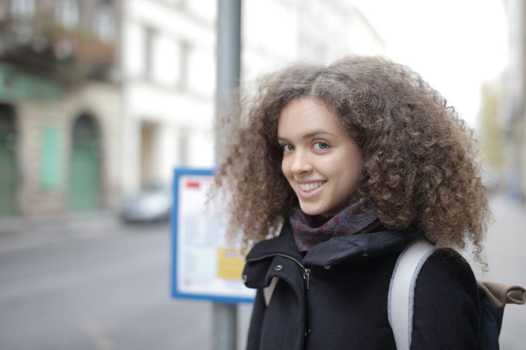 Positive female tourist wearing warm coat and smiling at camera while carrying backpack walking on city street in spring