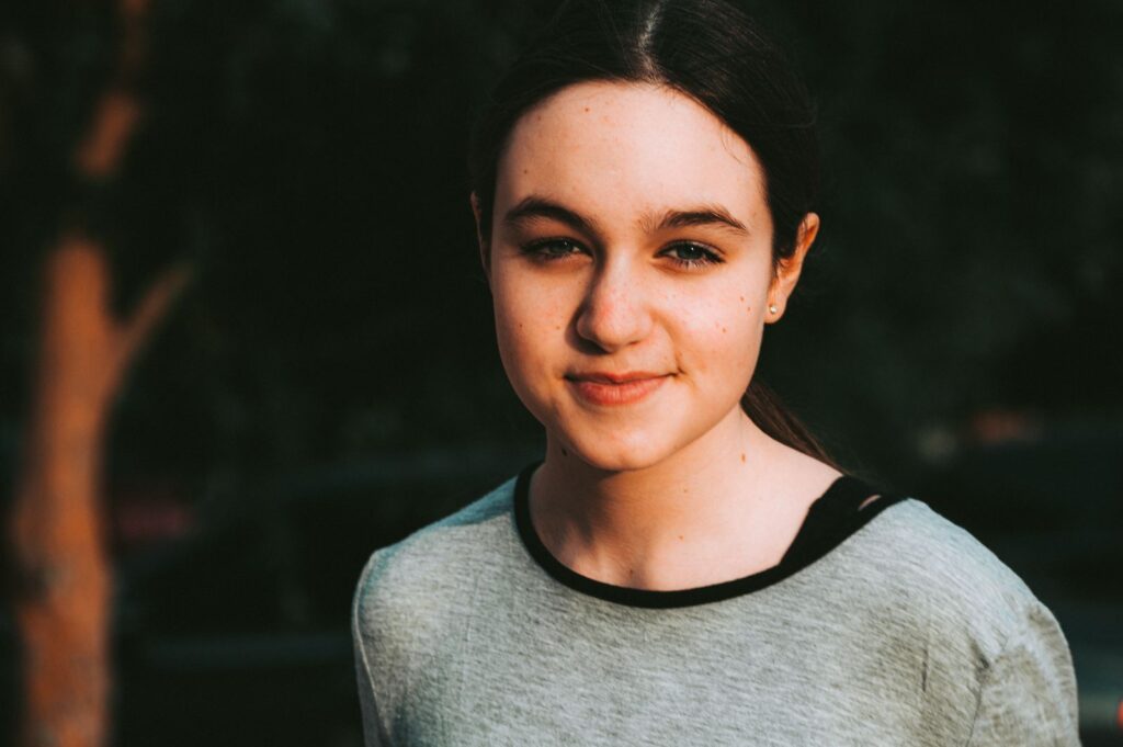 Pensive young female in grey shirt and dark hair tied up looking at camera in park on blurred background