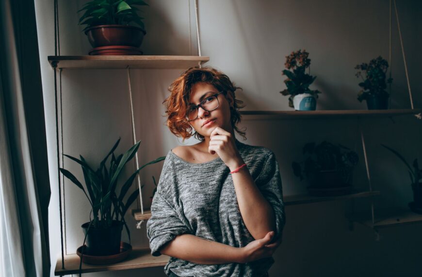 Young female in eyeglasses touching chin while thoughtfully looking at camera standing in room with plants on shelves