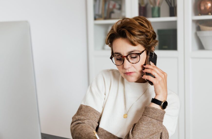 Good looking short haired female worker listening to work instructions on mobile and noting down information in modern office