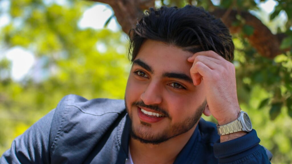 Happy young ethnic male in shirt with wristwatch touching forehead and looking away with bright toothy smile while resting in forest in daylight on blurred background